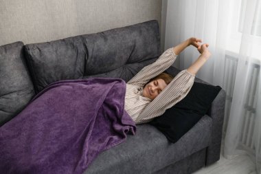 Gri bir kanepede uyandıktan sonra kollarını açan gülümseyen genç bir kadın, mor bir battaniyeyle kaplanmış, beyaz perdeli bir pencerenin yanında. İç mekan, doğal sabah ışığı..