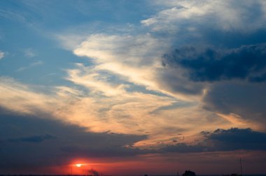 Yoğun bulutlu koyu mavi gökyüzü, yumuşak günbatımı ışığı ile aydınlanıyor, dramatik ve kasvetli bir atmosfer yaratıyor..