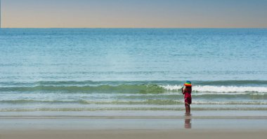 Kumsalda kum, deniz suyu ve sahilde yürüyen insanlar var.