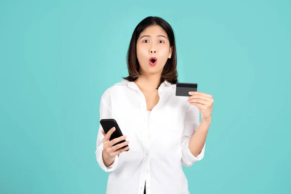Excited and surprised young asian woman standing isolated on bright green wall background studio. She using mobile cell phone and hold credit bank card. People lifestyle concept.