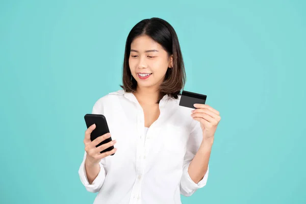 stock image Happy young asian woman standing isolated on bright green background. She using mobile cell phone and hold credit bank card. People lifestyle concept. Mock up copy space.