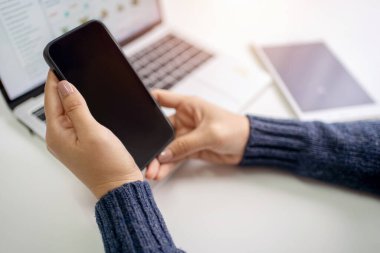 Kadın, boş ekranlı akıllı telefon ve yakınlardaki bir masada dizüstü bilgisayar kullanıyor. O, ofiste bir kazak giyerek oturuyor..