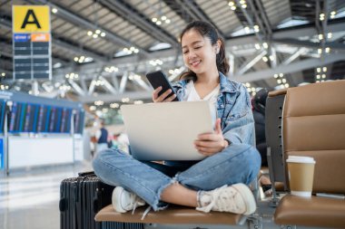 Pıhtılaşmış genç Asyalı kadın rahatla ve havaalanı terminalinde bavullu akıllı telefondan sosyal medya teknolojisini kullan..