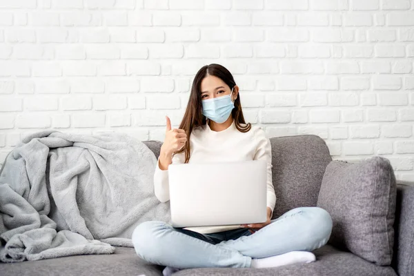 Asian woman in mask sits on sofa with laptop in living room.