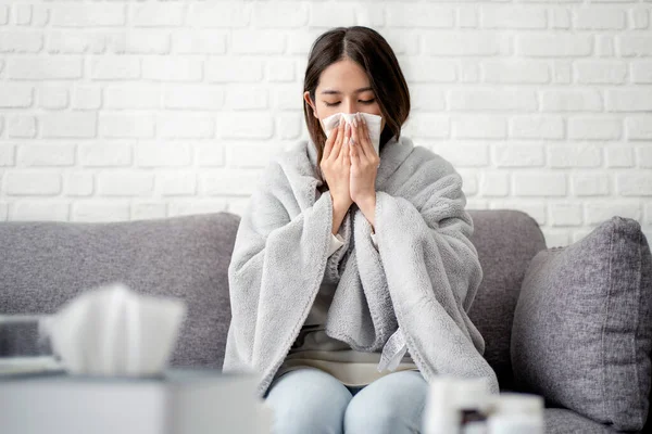 Sick Asian woman covered with blanket sneezing sitting on sofa She was sick and had a fever. influenza concept