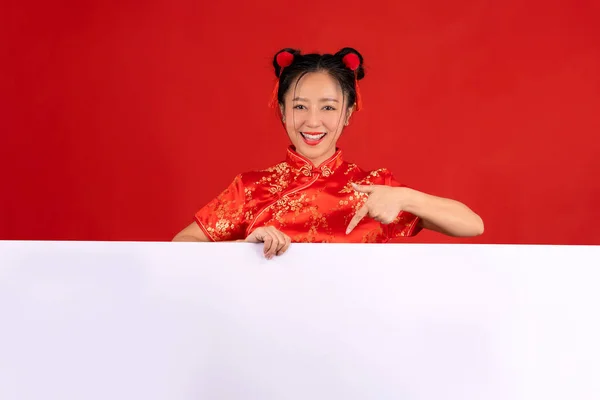 Happy asian woman in red casual attire with hand pointing blank announcement banner on red background. Happy Chinese new year.