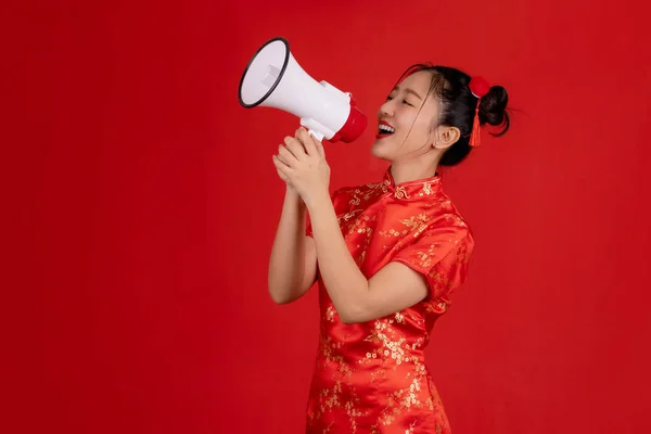 Happy Chinese New Year. Happy Asian woman wearing qipao is screaming with megaphone isolated on red background.