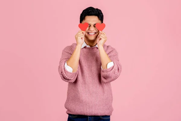 Young asian young man holding red love hearts over eyes isolated on pink background. Love and romantic concept.
