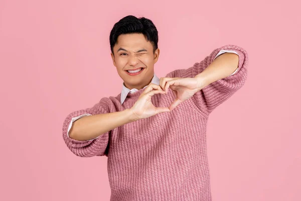 Asian handsome man in sweater standing on isolated pink background. He is smiling in love making a heart symbol with hands. Romantic concept