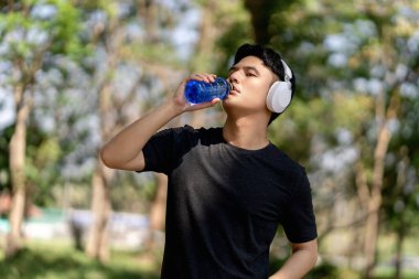 Erkek koşucu, kardiyo çalışmasından sonra plastik şişeden su içer. Sabah siyah spor bir takım elbise ve beyaz kulaklık takıyordu..