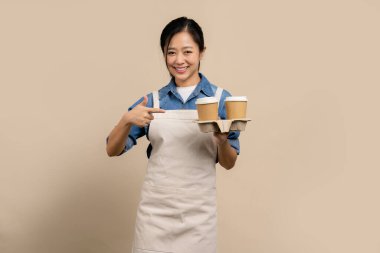 Kahve dükkanı, küçük işletme ve başlangıç konsepti. Neşeli Asyalı kadın barista krema rengi önlük giyer elindeki kahverengi kahve fincanını işaret eder..