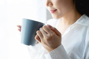 Çekici genç bir kadın beyaz gömleğiyle gülümsüyor sabahleyin pencerenin yanında otururken kahve içiyor. Boşluğu kopyala, kapat