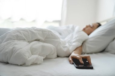 Güzel Asyalı kız beyaz yatakta uyuyor. Akıllı telefonu elindeyken uykuya daldı..