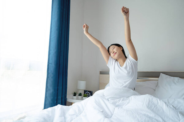 Asian beautiful girl in white nightgown wake up in the morning with happiness. She stretching body after getting up from sleep on bed in bedroom at home.