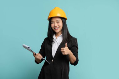 Asyalı kadın mühendis başparmağıyla ayakta duruyor ve izole edilmiş arkaplanda kontrol listesi tutuyor.