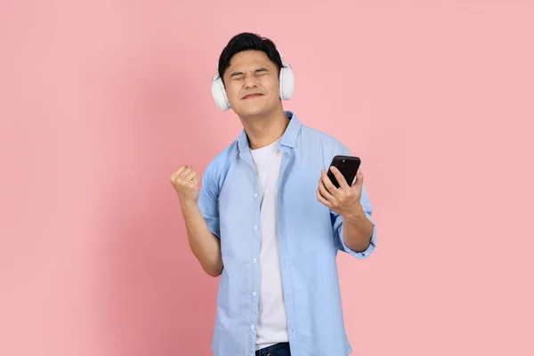 成功したアジアの男ハンサムな若い無線ヘッドフォンを身に着けているとピンクの空白のコピースペーススタジオの背景に隔離されたスマートフォンを使用して — ストック写真