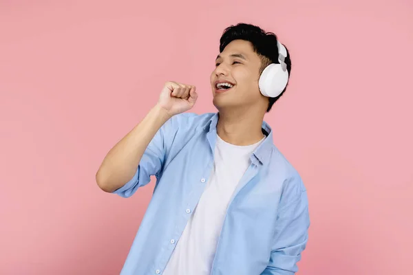 若いですハンサムなアジア人男性身に着けていますカジュアル服や無線ヘッドフォンながら歌彼のお気に入りの歌立って孤立した明るいピンクの背景 — ストック写真