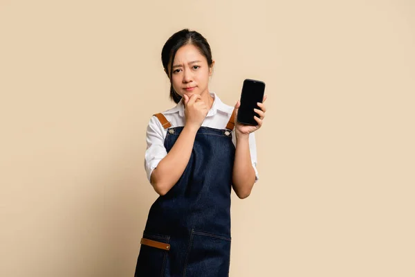 Unhappy young asian barista worker in apron holding smartphone with blank screen. She was wondering something and looked at the camera.