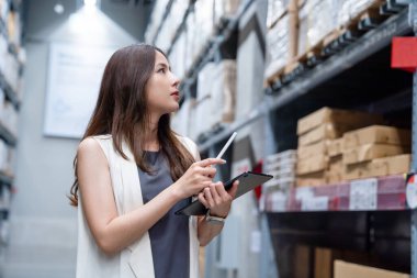 Genç zeki Asyalı iş kadını, fabrikadaki depoda ürün yönetimi için raflardaki malları kontrol etmek için dijital tablet kullanıyor.