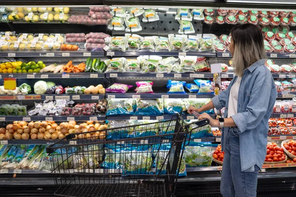 アジアの十代の少女がスーパーマーケットで買い物をする 彼女は棚に野菜を拾っている ストック画像