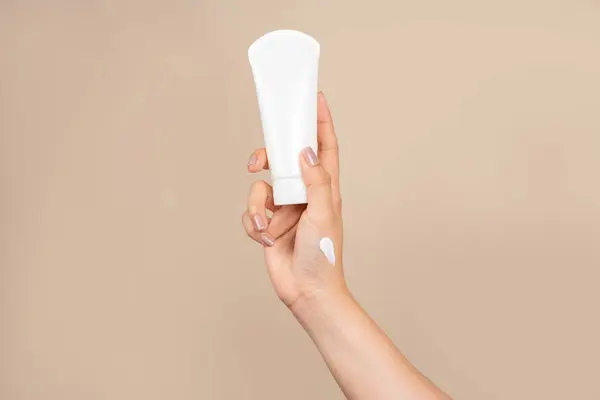 Female hand with cream to touch cosmetic tube on beige background. Concept of hand lotion cream or winter facial care.