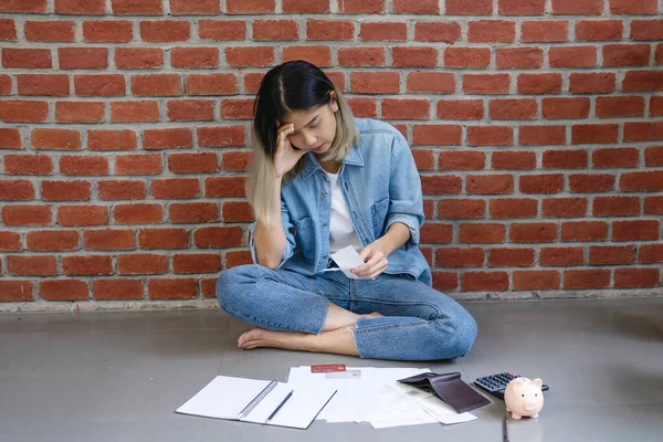 Stressed and confused asian woman sitting on floor home with calculating expense from invoice or bill , she has no money to pay.