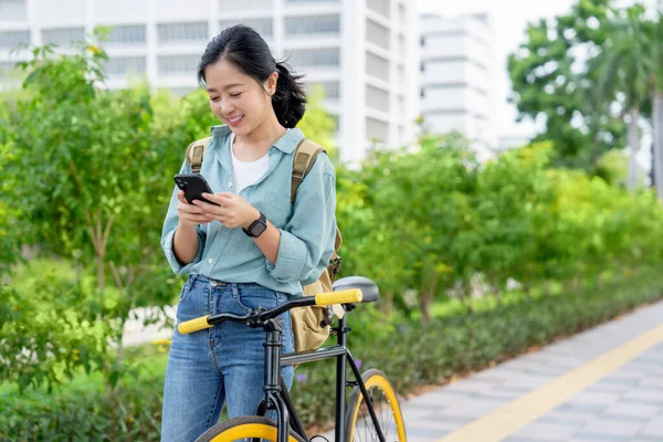 Happy young Asian woman in work clothes using smartphone with bicycle in outdoor park. Eco-friendly concept.