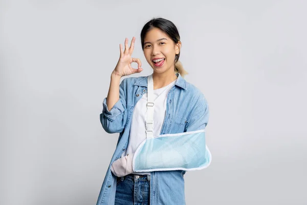 青いジーンズを着た美しいアジアのオフィスワーカーは 白い背景 生命および事故保険に大丈夫なサインを示す壊れた腕のために柔らかいスプリントを着用しました ロイヤリティフリーのストック画像