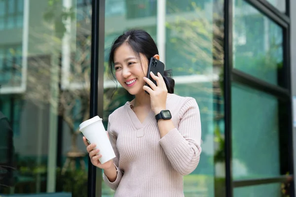 Relaxed Asian woman chatting on smartphone while walking in the city with a cup of coffee