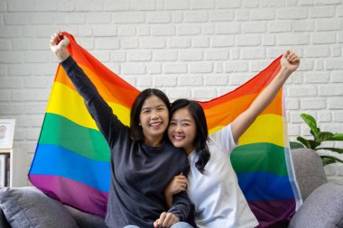 LGBTQ Onur Ayı, mutlu Asyalı lezbiyen çiftin portresi gökkuşağı bayrakları evdeki kanepede oturuyor.