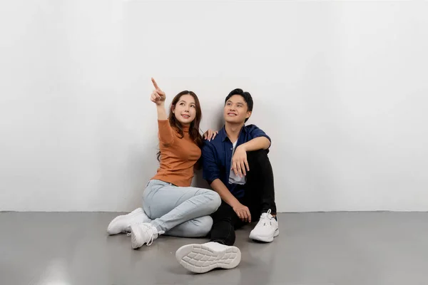 Portrait of cheerful young Asian couple in casual outfits sitting on floor and pointing at copy space