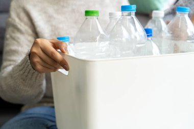 Krem rengi kazak giyen bir kadın oturma odasında geri dönüşümlü plastik şişeleri toplayıp beyaz çöp kutularına dolduruyor..