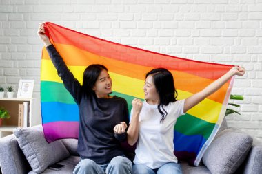 Mutlu genç Asyalı lezbiyen çift rahat bir kanepede oturup gülümsüyor ve ellerinde büyük bir gökkuşağı bayrağı tutuyorlar. Evdeki oturma odasında eşitlik için sevgi gösteriyorlar.