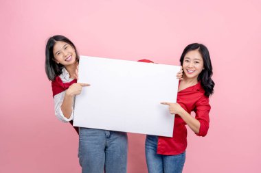 İki güzel Asyalı kadın pembe bir arka planda boş beyaz bir ilan panosunu gösterip tutuyorlar..