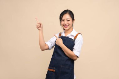 Önlüklü sevimli Asyalı kadın bej arka plandaki ilanınız için parmağını gösteriyor..