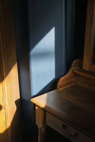 Güneş ışığının pencereden içeri aktığı, ahşap bir şifonyerin ve dolabın yanındaki dokulu mavi duvarın üzerine üçgen bir gölge çizdiği minimalist bir iç mekan..