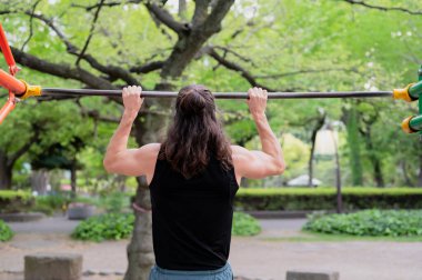 Kentteki yeşil yapraklı parktaki metal çubukta barfiks çeken kişi..