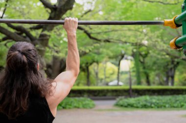 Kentteki yeşil yapraklı parktaki metal çubukta barfiks çeken kişi..