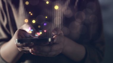 Yüzen sosyal medya simgeleriyle akıllı telefonu tutan ellerin yakın çekimi, dijital etkileşimi, gerçek zamanlı iletişimi ve modern teknoloji ve yaşam tarzı temaları için çevrimiçi kullanıcı katılımını sembolize ediyor.