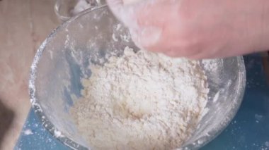 Women's hands add a chicken egg to the flour for making dough in a glass bowl.
