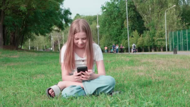 Glad Flicka Skolåldern Sitter Gräset Och Tittar Skärmen Sin Smartphone — Stockvideo
