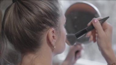 Kadın fırçayla makyaj yapıyor, dikkatlice aynaya bakıyor..