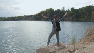 Woman tourist stands on top of mountain and, raising her hands, looks at seascape.