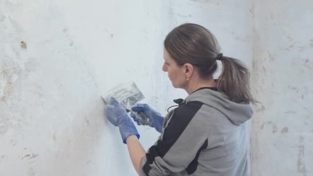 中年妇女正在整修她的公寓 她正在把旧石膏从墙上刮下来 — 图库视频影像