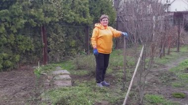 Turuncu kazaklı bir kadın bahçede elinde kürekle dikilip bahçe işlerinden zevk alıyor..