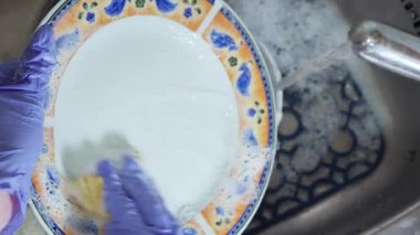Mavi eldivenli bir el, akan suyun altında köpüklü bir lavaboda süngerle tabağı yıkar..