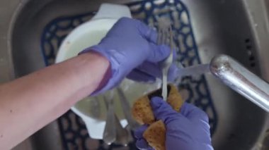 Mavi eldivenli bir kadın eli akan suyun altında süngerli bir çatal yıkar..