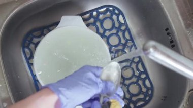 Mavi eldivenli bir kadın, köpüklü bir lavaboda bir kaşık suyun altında kaşık yıkıyor..