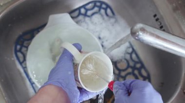 Mavi eldivenli bir el köpüklü lavaboda akan suyun altında bir fırçayla bardağı yıkar..