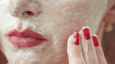 Bir kadın, dikkatli el hareketleri ve canlı kırmızı manikürle yüzüne yaşlanma önleyici krem sürüyor..